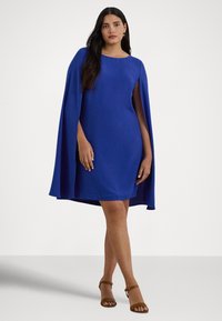 Modra oprijeta obleka z obliko, ki spominja na plašč, z okroglim izrezom in gladko teksturo. Kombinirana z rjavimi sandali z naramnicami.