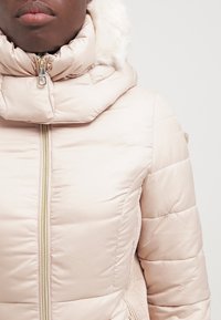 Beige prošívaná bunda s vysokým límcem, zapínáním na zip a umělou kožešinou na kapuci. Obsahuje texturované akcenty a přiléhavý střih.