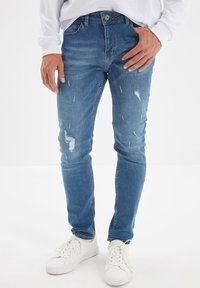Jean bleu en denim ajusté avec des zones usées et des détails de décoloration en forme de plis. Dispose d'un design traditionnel à cinq poches et d'une fermeture éclair.
