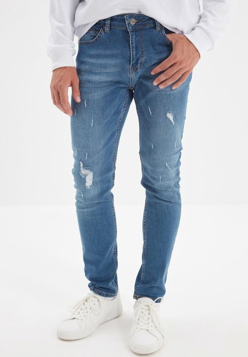 Jean bleu en denim ajusté avec des zones usées et des détails de décoloration en forme de plis. Dispose d'un design traditionnel à cinq poches et d'une fermeture éclair.