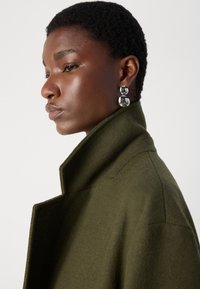 Olive green wool coat with a high collar. Model wears large metallic earrings, showcasing a smooth, shiny texture. Simple and modern design.