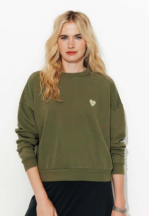 Sweatshirt vert olive avec un col ras du cou, design à épaules tombantes et un patch en forme de cœur texturé sur la poitrine gauche. Poignets et ourlet côtelés.