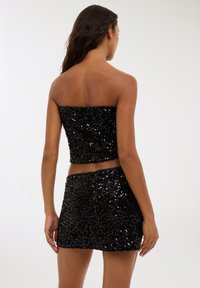 Kobieta z długimi włosami ubrana w czarny top bez ramion z cekinami i dopasowaną mini spódnicą, odwrócona plecami na jednolitym jasnym tle.