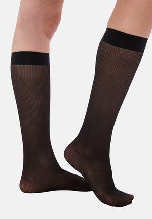 Beine, die schwarze, halbtransparente, knielange Strümpfe mit geripptem Muster und solid schwarzen Bändern am oberen Rand tragen, vor einem weißen Hintergrund posieren.