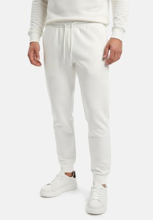 Uomo che indossa joggers bianchi aderenti con vita a coulisse e sneakers bianche con dettaglio nero sul tallone, in piedi su sfondo semplice.