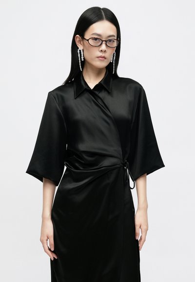 Vestido envolvente de satin preto com gola, mangas ¾ e um cinto atado na cintura. Textura suave com um design drapeado. Brincos de declaração claros.