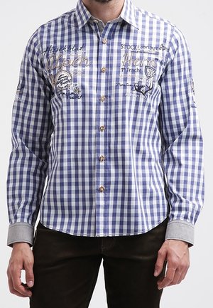 Mann trägt ein blau-weiß kariertes Hemd mit Knöpfen, besticktem Text und bergthematischen Motiven auf Brust und Ärmeln.