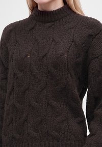 Gros plan d'une personne portant un pull épais en maille marron foncé avec un col montant côtelé et un motif torsadé.