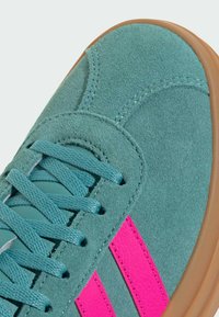 Turkos mockasinsneaker med rosa ränder, med teal snörning och en gummisula. Närbilden framhäver textur och sömdetaljer.