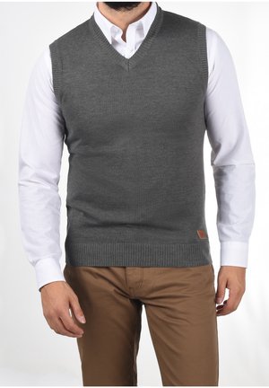 Gilet gris en tricot avec un col en V, ourlet côtelé, porté sur une chemise blanche, assorti à un pantalon marron. Présente un petit détail en cuir avec un logo.