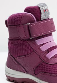 Bota infantil de media altura en color púrpura, fabricada en material sintético texturizado, con una tira de velcro y cordones elásticos, y suela de goma.