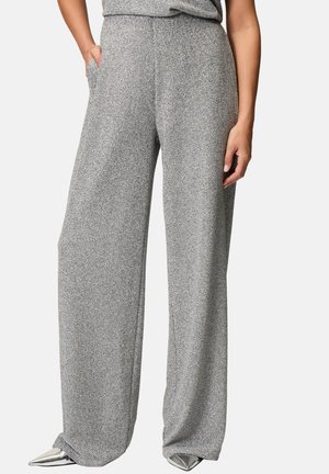 Femme portant un pantalon gris texturé taille haute à jambes larges, associé à des chaussures argentées à bout pointu, une main dans la poche.