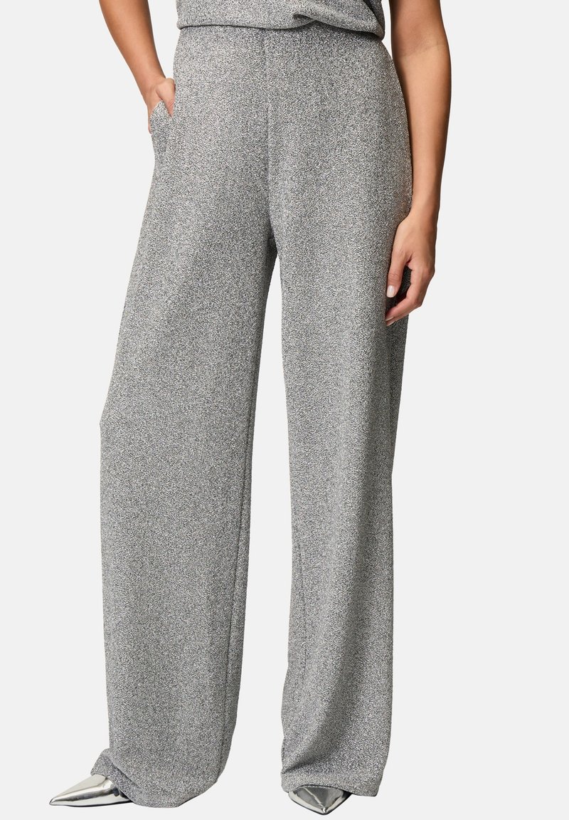 Femme portant un pantalon gris texturé taille haute à jambes larges, associé à des chaussures argentées à bout pointu, une main dans la poche.
