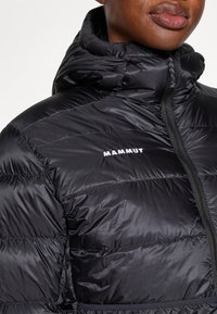 Juodas pūkinis švarkas su aukštu apykakle, su priekiniu užtrauktuku ir horizontaliais dygsniavimais. Prekės ženklas „MAMMUT“ pateiktas baltu rašalu.
