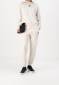 Sudadera beige con logotipo negro "HUGO", combinada con pantalones de chándal a juego. Zapatillas negras con suelas blancas. Tejido texturizado, corte relajado.