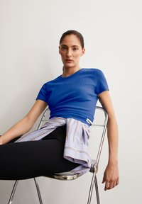 Blauwe kortärmige, getailleerde top met ribbelstructuur, gecombineerd met een gestrikt gestreept shirt. Zwarte leggings en een chroomstoel voor een neutrale achtergrond.