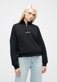 Νέα γυναίκα με μακριά ξανθά μαλλιά, φορώντας μαύρο φούτερ με μισή ζώνη και το κείμενο "NAPAPIJRI" και ανοιχτόχρωμο τζιν, στέκεται μπροστά σε λευκό φόντο.