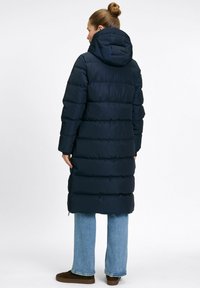 Vrouw die met haar rug naar de camera staat, gekleed in een lange marineblauwe pufferjas met capuchon, lichte spijkerbroek en bruine schoenen, tegen een effen witte achtergrond.