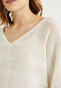 Neulepaita, jossa on v-pääntie, kerma väri ja avonainen kudontamalli raitatekstuurilla. Pehmeä kangas, jossa on vuorottelevat tiukat ja löysät neuleet.