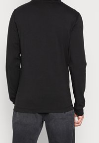 Person, der ein schlichtes schwarzes Langarmshirt und dunkelgraue Jeans trägt, von hinten vor einem einfarbigen Hintergrund gezeigt.