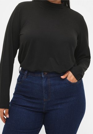 Femme portant un col roulé noir à manches longues rentré dans un jean bleu foncé taille haute, avec une main dans la poche.