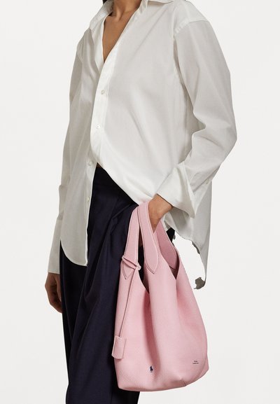Ροζ δερμάτινη τσάντα με υφή, διαθέτοντας διπλούς καμπύλους ιμάντες και μια μικρή ετικέτα. Φορεμένη με λευκό πουκάμισο και σκούρους παντελόνες.