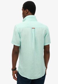 Uomo che indossa una camicia di lino a maniche corte azzurro chiaro e pantaloni scuri, visto da dietro su sfondo bianco semplice.