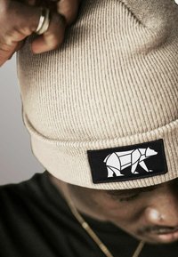 Man adjusting beige knit beanie with black patch featuring a white geometric bear design, wearing a black shirt and gold chain.