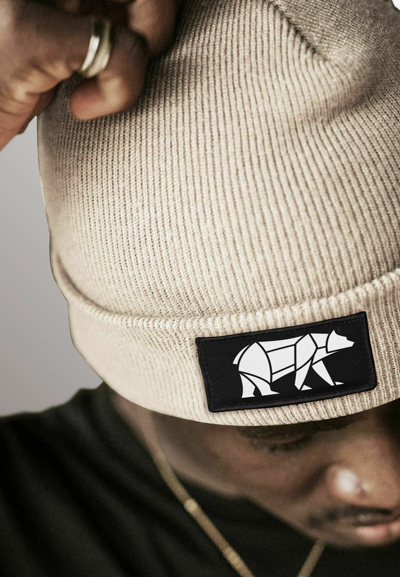 Man adjusting beige knit beanie with black patch featuring a white geometric bear design, wearing a black shirt and gold chain.