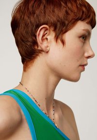 Top asimétrico verde y azul en una modelo, con cabello rojo corto y un collar de cuentas multicolores, que incluye un delicado acento dorado.
