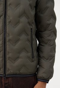 Giacca puffer verde oliva con motivo a chevron, zip frontale e tasche laterali. Presenta polsini elastici e orlo nero con cuciture a contrasto.