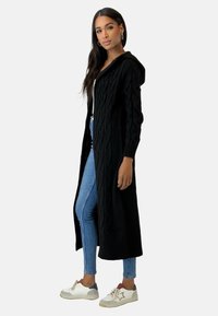 Svart, lång stickad cardigan med huva, med kabelmönster och ribbade ärmslut, parat med ljusblå jeans och vita sneakers.