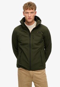 Veste softshell vert foncé avec fermeture éclair à l'avant et capuche. Comprend une poche poitrine et deux poches latérales. Portée par-dessus un pull de couleur claire.