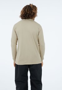 Beżowa koszula z długim rękawem o gładkiej fakturze i zaokrąglonym dole, noszona z czarnymi spodniami. Tył ma prosty i nieozdobiony design.