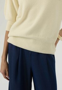 Personne portant un pull en maille crème avec des poignets et un ourlet côtelés, associé à un pantalon bleu foncé ample, main posée sur la hanche.