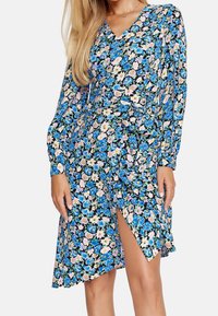 Modelka w długiej sukience z długim rękawem w kwiatowy wzór, z niebieskimi i różowymi kwiatami, z wysokim rozcięciem na udzie, na jednolitym tle.