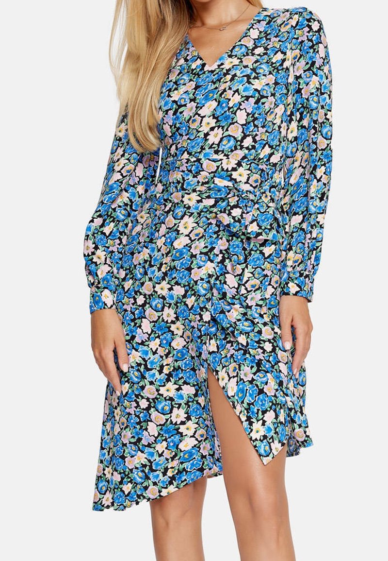 Modelka w długiej sukience z długim rękawem w kwiatowy wzór, z niebieskimi i różowymi kwiatami, z wysokim rozcięciem na udzie, na jednolitym tle.