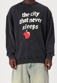 Sudadera gris oscuro con cuello redondo, que presenta un texto blanco que dice "la ciudad que nunca duerme" y un gráfico de una manzana roja con un mordisco.