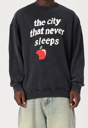 Sudadera gris oscuro con cuello redondo, que presenta un texto blanco que dice "la ciudad que nunca duerme" y un gráfico de una manzana roja con un mordisco.