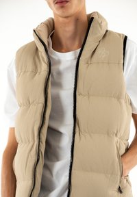 Chaleco acolchado beige con textura acanalada, cierre de cremallera y cuello alto. Llevado sobre una camiseta blanca, que presenta un logo bordado en el pecho.