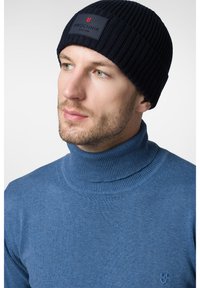 Granatowa, prążkowana czapka z logo oraz niebieski sweter z golfem o subtelnej fakturze. Zbliżenie na głowę i górną część ciała.