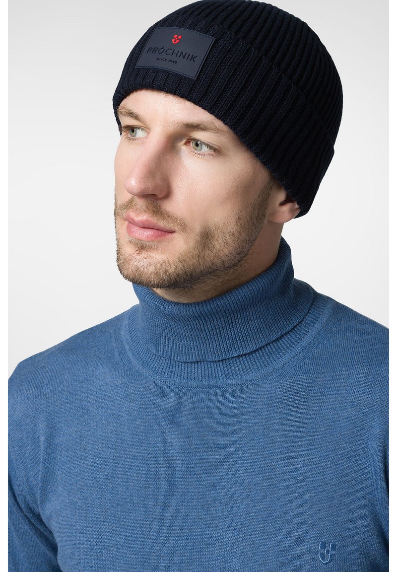 Granatowa, prążkowana czapka z logo oraz niebieski sweter z golfem o subtelnej fakturze. Zbliżenie na głowę i górną część ciała.