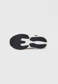 Semelle de chaussure de sport noire et blanche avec un design en caoutchouc texturé, présentant des rainures de traction bien définies et une zone de talon arrondie.