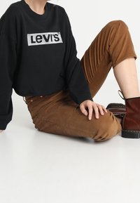 Musta lyhyt collegepaita hopeisella "Levi's"-logolla, yhdistettynä ruskeisiin samettihousuihin ja ruskeisiin nilkkureihin. Sileä pinta ja rento istuvuus.
