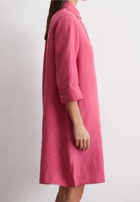 Kobieta w różowej lnianej sukience o długości do kolan, z długim rękawem i kołnierzykiem, stojąca bokiem na jednolitym jasnym tle.