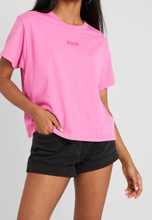 Vrouw draagt een felroze Levi's T-shirt en zwarte opgerolde denim shorts, staand met één hand in de zak tegen een grijze achtergrond.