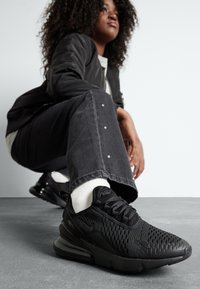 Juodi Nike sportbačiai su tinkline viršutine dalimi, vėdinimo detalėmis, tekstūruotu padu ir paminkštintu apykakle, derinami su pilkais džinsais ir baltais kojiniais.