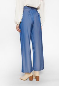 Wijdvallende broek in textuurblauwe stof, met een hoge taille en plooien. Gepaarde met lichtbeige enkellaarsjes.