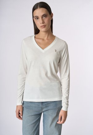 T-shirt bianco a maniche lunghe e scollo a V leggero, con un logo sottile sul lato sinistro del petto, indossato con pantaloni in denim blu. Texture liscia.