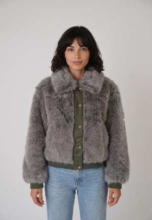 Femme aux cheveux foncés portant une veste en fausse fourrure grise par-dessus un haut vert boutonné et un jean bleu clair, debout devant un fond uni.